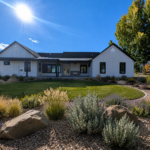 Modern farmhouse backyard in Cache Valley, Utah with white board-and-batten siding, black trim, covered patio, outdoor fireplace, green lawn, and Utah native landscaping with ornamental grasses, shrubs, gravel, and boulders.