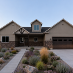 Front exterior of Carpenter Custom Home in Cache Valley, Utah with tan siding, brick accents, covered entry, dark garage door, and concrete driveway.